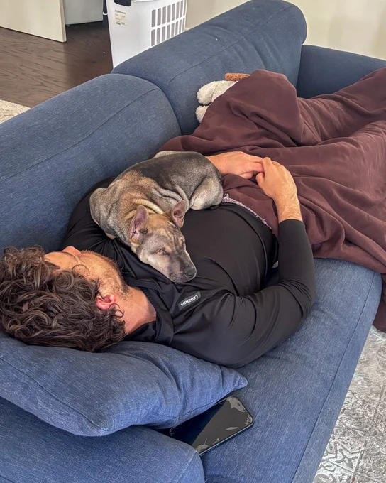 Image of Tyson Jost taking a nap on a couch with his new foster puppy Linguine sleeping on his chest 