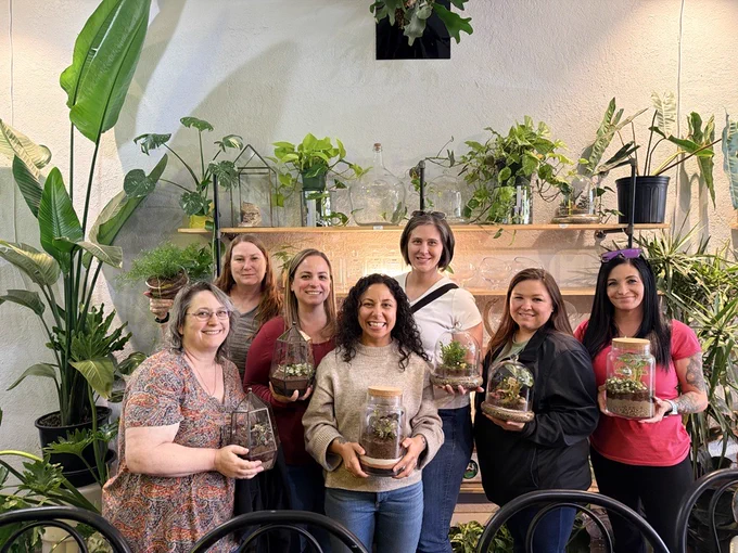 Group of seven people holding glass terrariums filled with plants, standing in a room decorated with various green potted plants on shelves and floor.