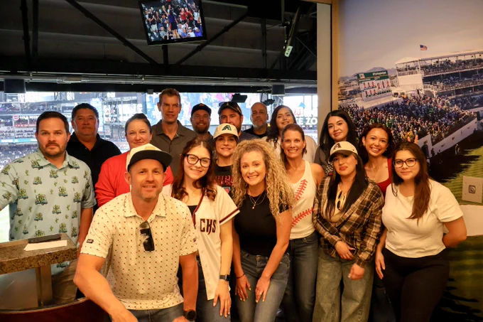 Group of people posing together inside a sports stadium suite with a baseball game visible on TV above them.