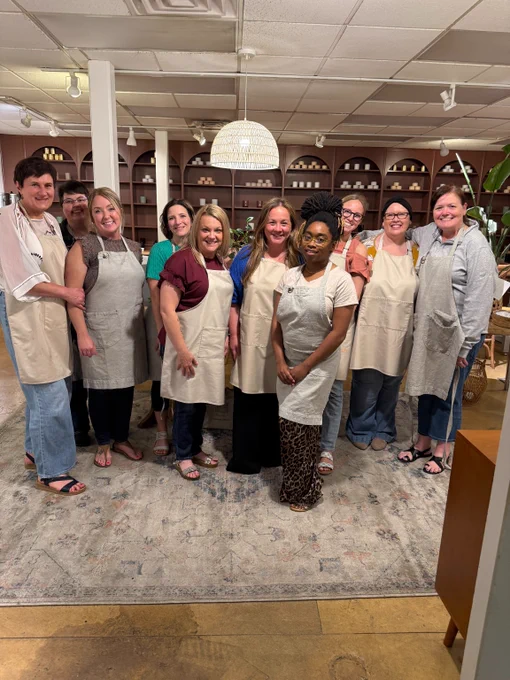 Group of nine people wearing aprons standing together in a cozy room with shelves of candles behind them.