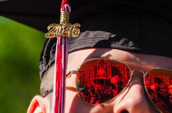 Close-up of a Ball State graduate’s cap, 2026 tassel, and sunglasses reflecting the Spring Commencement ceremony and campus buildings.