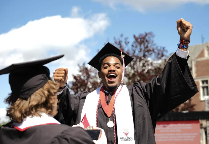 A Ball State graduate in cap and gown celebrates with raised fists and a wide smile during Spring Commencement, with another graduate in the foreground.