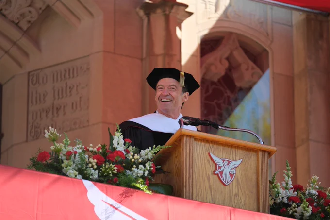 Hugh Jackman smiles while speaking at a podium during Ball State University’s Spring Commencement ceremony, wearing academic regalia in front of campus architecture and floral arrangements.