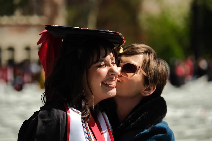 A Ball State graduate in cap and gown smiles as a loved one kisses their cheek during Spring Commencement celebrations outdoors.