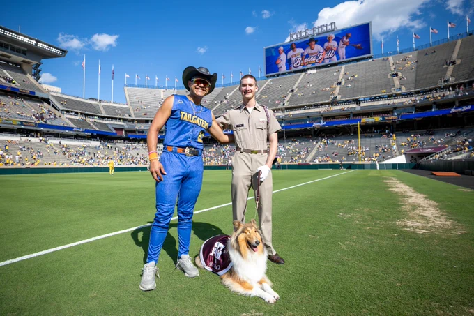 A person in a blue "Tailgaters" uniform poses with another individual in a uniform on a sports field, accompanied by Texas A&M mascot, Reveille, wearing a Texas A&M blanket. The stadium is sparsely filled, and a scoreboard displays "Howdy!" with images of Banana Ball players.
