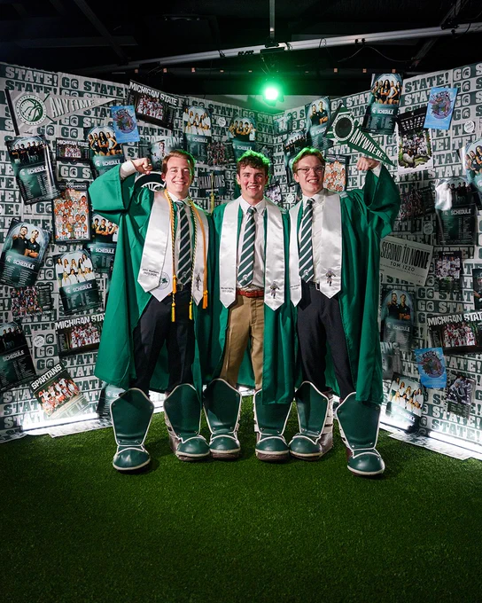 Three students in graduation gowns stand on artificial turf in front of a wall covered in Michigan State University memorabilia. They wear green gowns and white stoles and commemorative green and white Sparty boots.