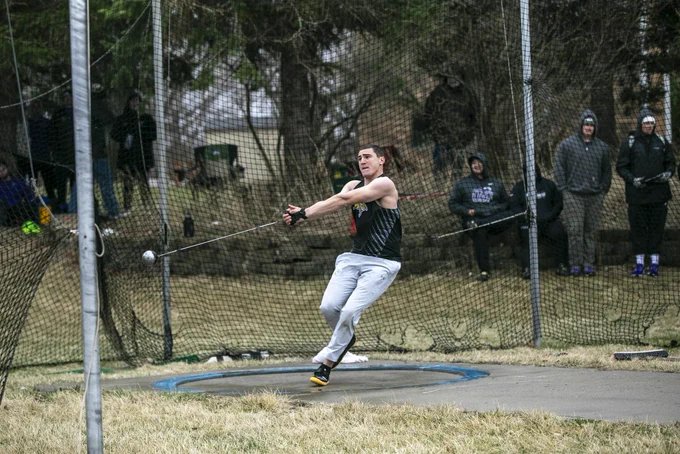A male track athlete in a black uniform top and grey sweat pants spins around in the throwing pit, beginning his hammer throw attempt
