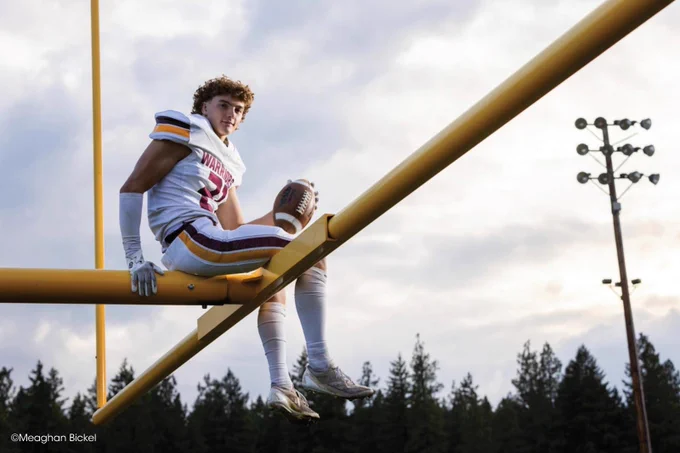A football player in a "Warriors" uniform is seated on a yellow goalpost, holding a football under one arm. The sky is cloudy, and trees are visible in the background. Copyright Meaghan Bickel.