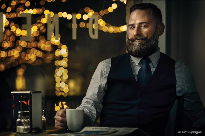 A well-dressed individual sitting at a table in a dimly lit cafe with a warm ambiance. They are holding a cup, and there's a newspaper on the table with blurry, festive lights in the background. Copyright Curtis Sprague.
