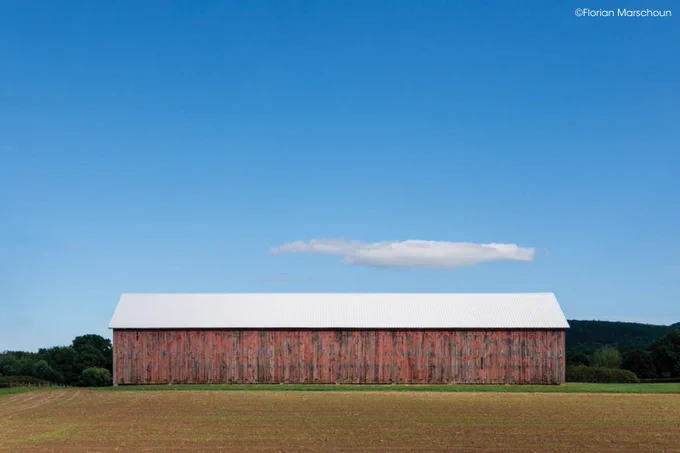 A large red barn with a white roof stands in a field under a clear blue sky. A single cloud floats above the barn. Copyright Florian Marschoun.