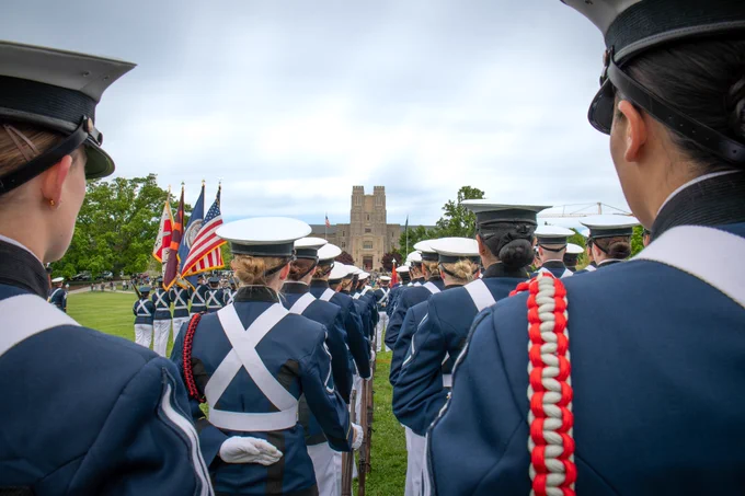 View from behind cadets standing at parade rest in formation, facing the Drillfield with flags and campus buildings in the distance.
