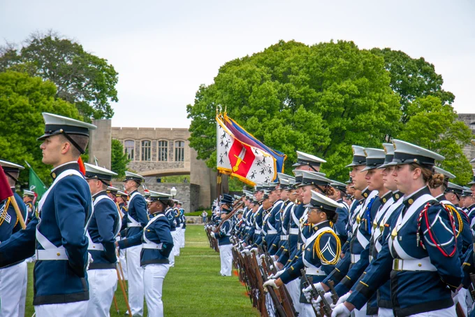 Cadets stand in formation at parade rest on the Drillfield, with multiple unit flags displayed behind them.
