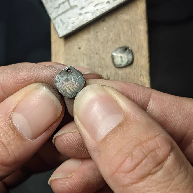 Two small meteorite charms with faint etched patterns: one held between fingertips, the second resting on a wooden bench pin in the background.