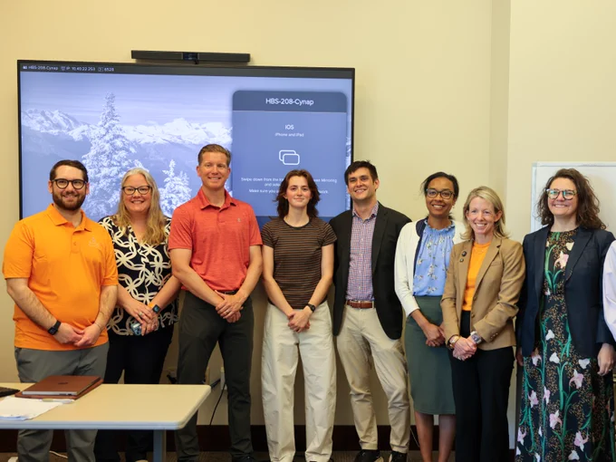A group of eight people are standing indoors, smiling at the camera. They are positioned in front of a large screen with an image of snowy mountains and trees. The group is dressed in business casual attire.