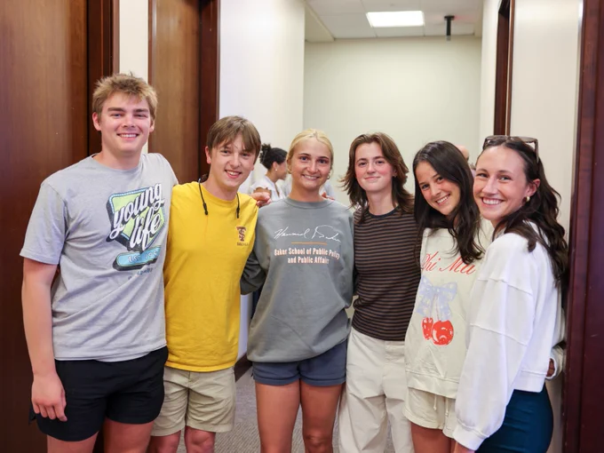 A group of six young people stands together in a hallway, smiling at the camera. They are casually dressed in t-shirts and shorts.