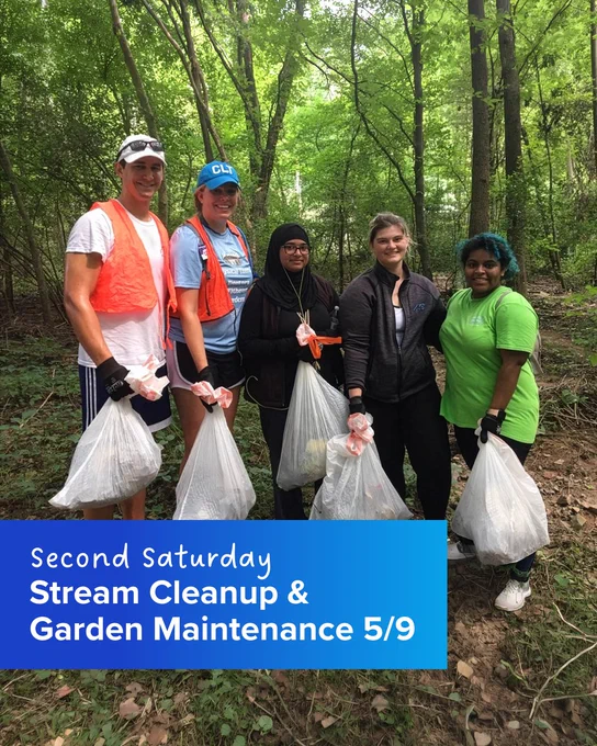 A group of five people stands in a forested area, holding bags filled with collected trash. They are participating in an event titled "Second Saturday Stream Cleanup & Garden Maintenance 5/9," as indicated by a text overlay. The surroundings are lush with trees and greenery.