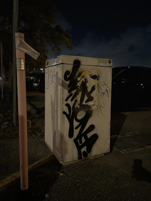 Nighttime view of a beige utility box covered in black graffiti. Next to it, a label is peeling off a safety bollard. The scene reflects infrastructure neglect in a dimly lit urban area.