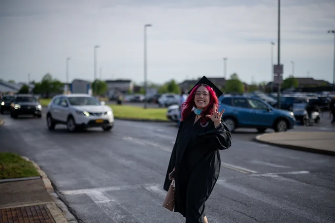 A graduate with pink hair and glasses wears a black gown, waving and smiling while crossing a street with cars and buildings in the background.