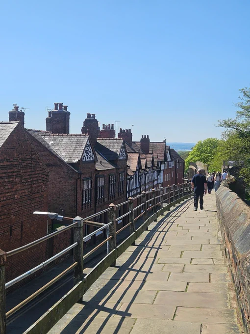 Sunny day with a clear blue sky; people walking on a stone city wall with a metal fence to the left. Red brick terraced houses on the left hand side and green trees to the right. In the distance, hills can be seen. 