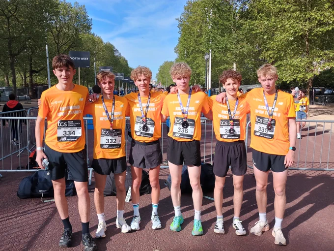 Six young male runners in orange t-shirts and medals pose together after a race.
