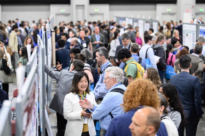 A large group of Annual Meeting attendees engaged in discussions around numerous poster displays in a spacious Poster Hall. Attendees are dressed in business attire. The atmosphere is bustling and collaborative.