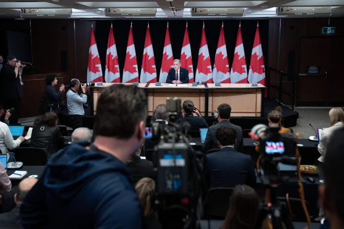 Prime Minister Mark Carney sits at a press conference table and speaks to reporters with cameras pointed toward him.