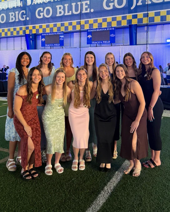 11 women dressed in semi-formal attire pose for a team photo in an indoor athletic facility