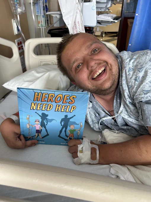 A young man with a partially shaved head and fresh surgical wound is smiling in his hospital bed as he holds a copy of his HEROES NEED HELP. The picture book features two children, each attached to an IV pole. Their shadows are depicted as super heroes.