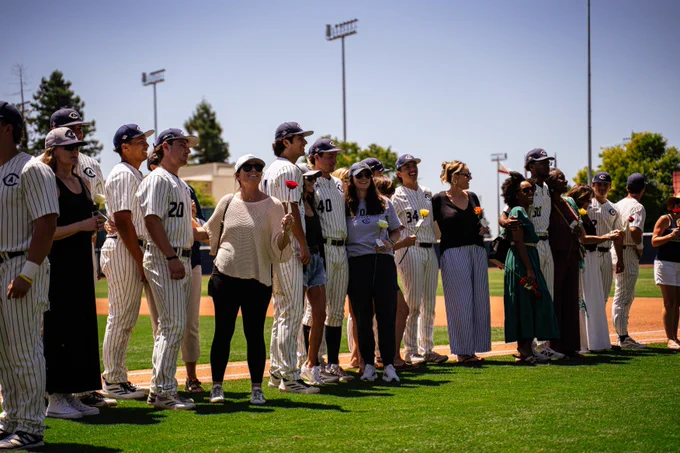 Players and their moms line up along the foul line, roses in hand, under a bright blue sky.