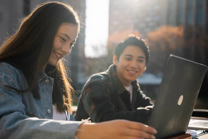 Two teenagers studying outdoors, smiling while using a laptop in sunlight.