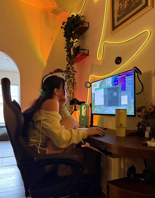 indie game dev at her desk in a hoodie, green ambient lights and plants on the walls, monitor full of code youre not allowed to see. building DA GAME and testing agents on the side
