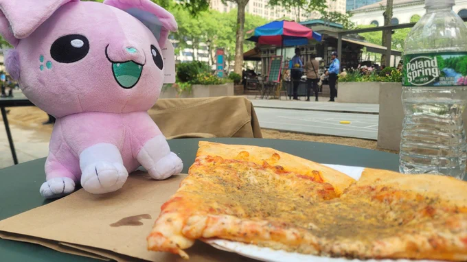 Mutt plushie from TopDog Studios in a park setting in New York City accompanied by 2 slices of pizza