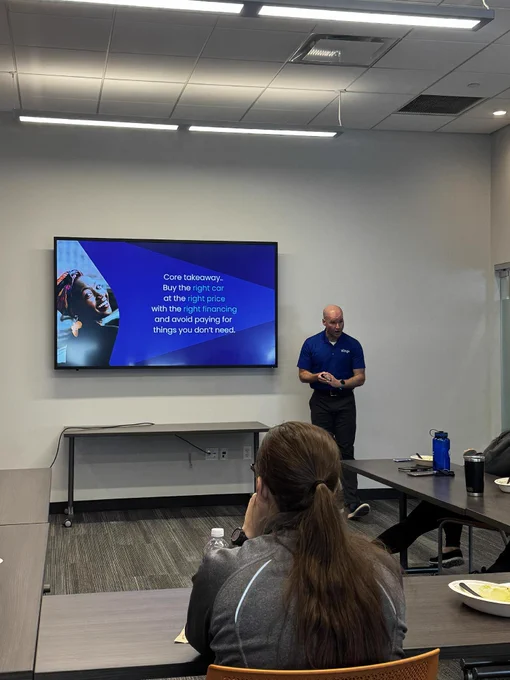 A person stands in front of a screen presenting a slide in a conference room. The slide advises, “Core takeaway: Buy the right car at the right price with the right financing and avoid paying for things you don’t need.” Two people sit at the table, listening.