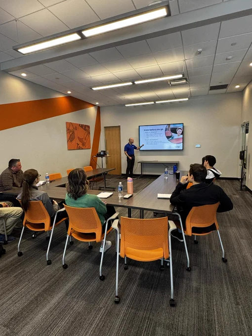 A person is giving a presentation in a conference room with seven others seated at a table. A slide on the screen is titled "Tesla Buckle up and get ready!" The room has orange accents, and a geometric painting is displayed on the wall.