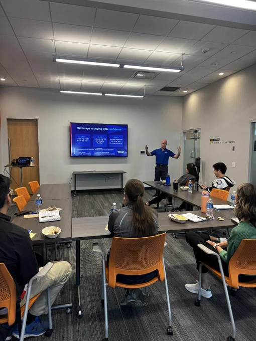A person is giving a presentation in a conference room to a group of seated attendees. The presentation slides are displayed on a wall-mounted monitor. The room has a U-shaped table arrangement and modern lighting.