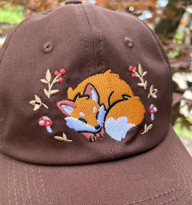 A brown hat featuring an embroidered fox design. The fox is sleeping with its fluffy tail curled next to its face. It is surrounded by little plants and mushrooms.