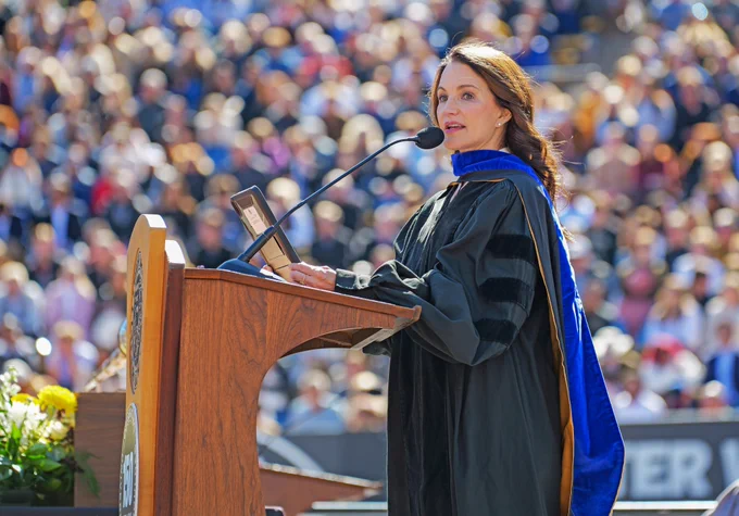 Kristin Davis stands at a podium wearing black graduation regalia and a blue and gold hood. Credit: University of Colorado