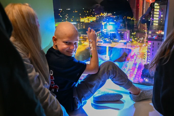 A patient at Children's Hospital of Richmond at VCU is seated by a window and smiling while looking out at a cityscape illuminated with colorful lights from emergency vehicles from around the Richmond region participating in the fourth annual goodnight lights event.