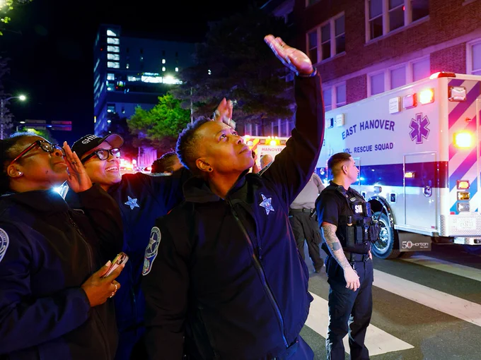 Public safety personnel are gathered on a street at night, looking upwards at Children's Hospital of Richmond. Emergency vehicles, including one marked "East Hanover, Medical Rescue Squad," are in the background with flashing lights during the fourth annual goodnight lights event.