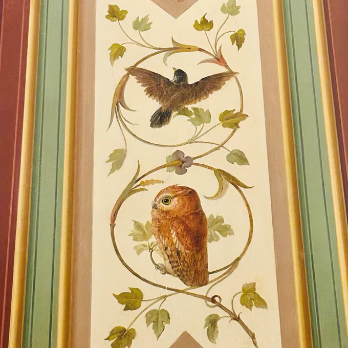 A detailed mural depicting a brown owl perched on a vine with leaves, along with a flying bird above, set against a decorative background with shades of maroon, green, and beige.