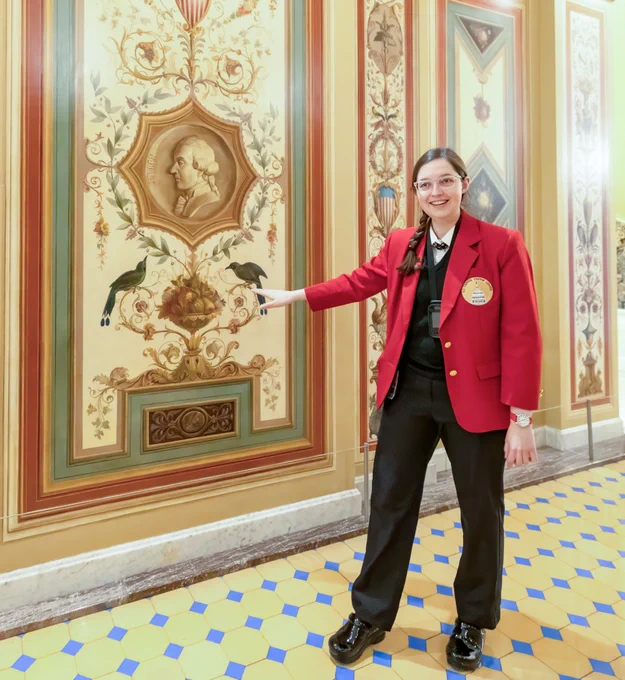 A tour guide wearing a red jacket stands indoors, pointing at an ornate wall mural featuring detailed artwork and a portrait in a decorative frame. They wear a lanyard and badge, and the floor has a yellow and blue geometric tile pattern.