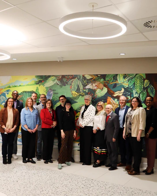 A group of people standing in front of a colorful wall mural depicting a vibrant nature scene at the Robert Bateman Community Centre in Burlington, Ontario. They are in an indoor setting with circular ceiling lights.