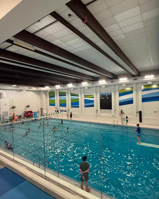 Centennial Pool in Burlington, Ontario with several individuals swimming and others standing by the poolside. The pool is bordered by a glass barrier. The facility features high ceilings with beams and walls decorated with blue, green, and white wave patterns.