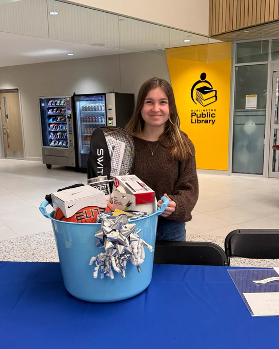 A person stands behind a table, smiling, in a public library. In front of them is a blue bucket filled with various items, including a tennis racket and a basketball. The yellow sign in the background reads "Burlington Public Library."
