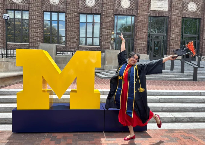Joyful student throwing up her arms next to a giant block M in front of the Hatcher Library.