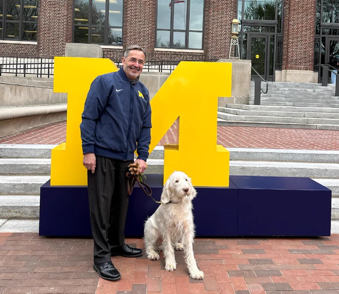 U-M President Grasso and his adorable dog standing in front of a giant block M.