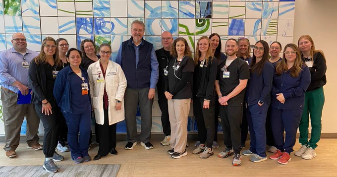 A group of about fifteen medical professionals stand smiling in front of a blue and green abstract tiled wall. Some are wearing scrubs, others lab coats, and one person is in a vest.