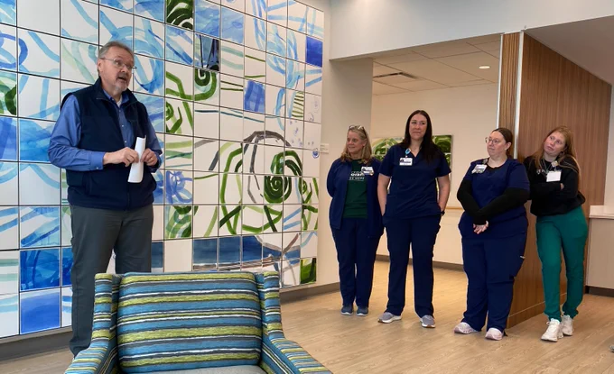 Man speaking to four women in scrubs in front of wall art with green and blue designs. The women are listening to the man, standing in a row. There is a chair in front of the wall art.
