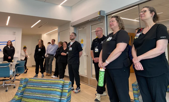 A group of healthcare workers in scrubs are standing and looking to their left. Some are wearing eyeglasses. A woman holds a green water bottle with white floral patterns. The setting appears to be a modern medical facility.