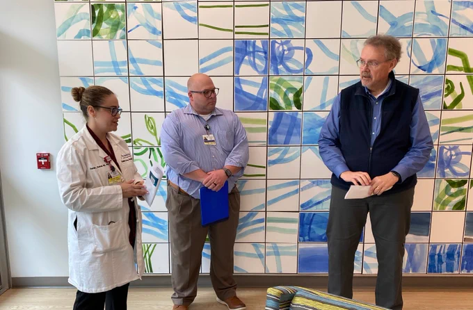 Three people converse in front of a wall with blue and green abstract art. On the left is a woman in a white lab coat with an ID. Next to her, a man holds a blue folder. A third man stands with a paper in hand.